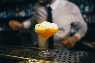 Close-up of an egg white cocktail sitting on the bar. In the background is a bartender wearing a tie.