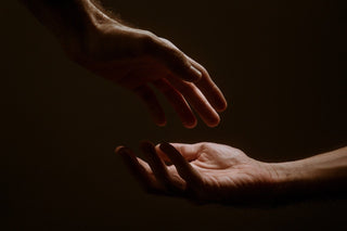 Two hands slightly in the dark about to touch. It looks like the hands are reaching out to each other in the act of service or to provide support. 