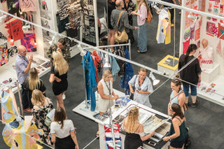 Bird's eye view of an exhibition show. Groups of people are mingling around various trade show display booths displaying various products including clothes, accessories and jewlery.