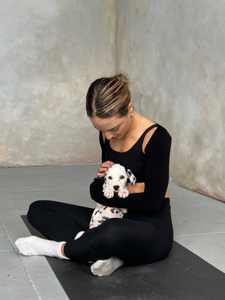 A woman in an an all-black athletic outfit is partially holding up a Dalmation puppy in the crook of her arm. The woman is sitting cross-legged on a black yoga mat. She is petting the Dalmation gently on its head. 