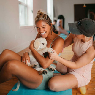 A seated woman in matching teal shorts and sports bra is holding a white labarador puppy in her arms while seated on a teal yoga mat. Behind her is another women with her face covered by a black baseball cap (in pink athletic unitard). The woman in the black cap is holding on the white labrador puppy's paw. In front of a the black capped woman is a glass of mimosa - as part of the puppy yoga and bubbly event