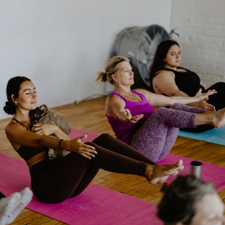Three women in the middle of a seated yoga pose. The women in matching brown yoga set is holding a puppy in her arms during a puppy yoga and bubbly class. 