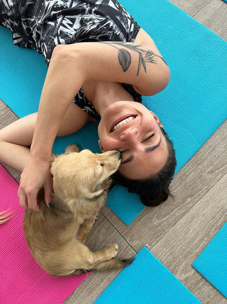 Woman lying down on a teal mat while a small puppy is sitting by her face giving her kisses during a puppy yoga class