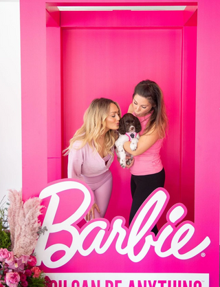 Two women wearing pink and holding a puppy are standing in a life-sized Barbie-packaging photo op