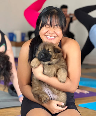 Woman laughing hard while holding a sleeping puppy chow chow in her arms. The woman is kneeling on a printed purple yoga mat. In the background are various people in mid-flow during a puppy yoga & bubbly event featuring chow chows. 