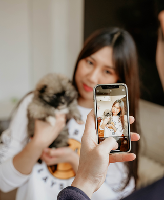 A woman is holding up a pomsky and posing for a camera photo while an arm holding an iphone is in frame about to take an image of the woman and puppy during a puppy yoga & bubbly event. 