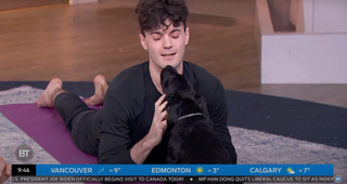 A screencap of a live broadcast on Breakfast Television to brought puppy yoga & bubbly to the Breakfast Television studio. In the screencap is a man lying on a purple yoga mat gently pet a black labrador puppy. 